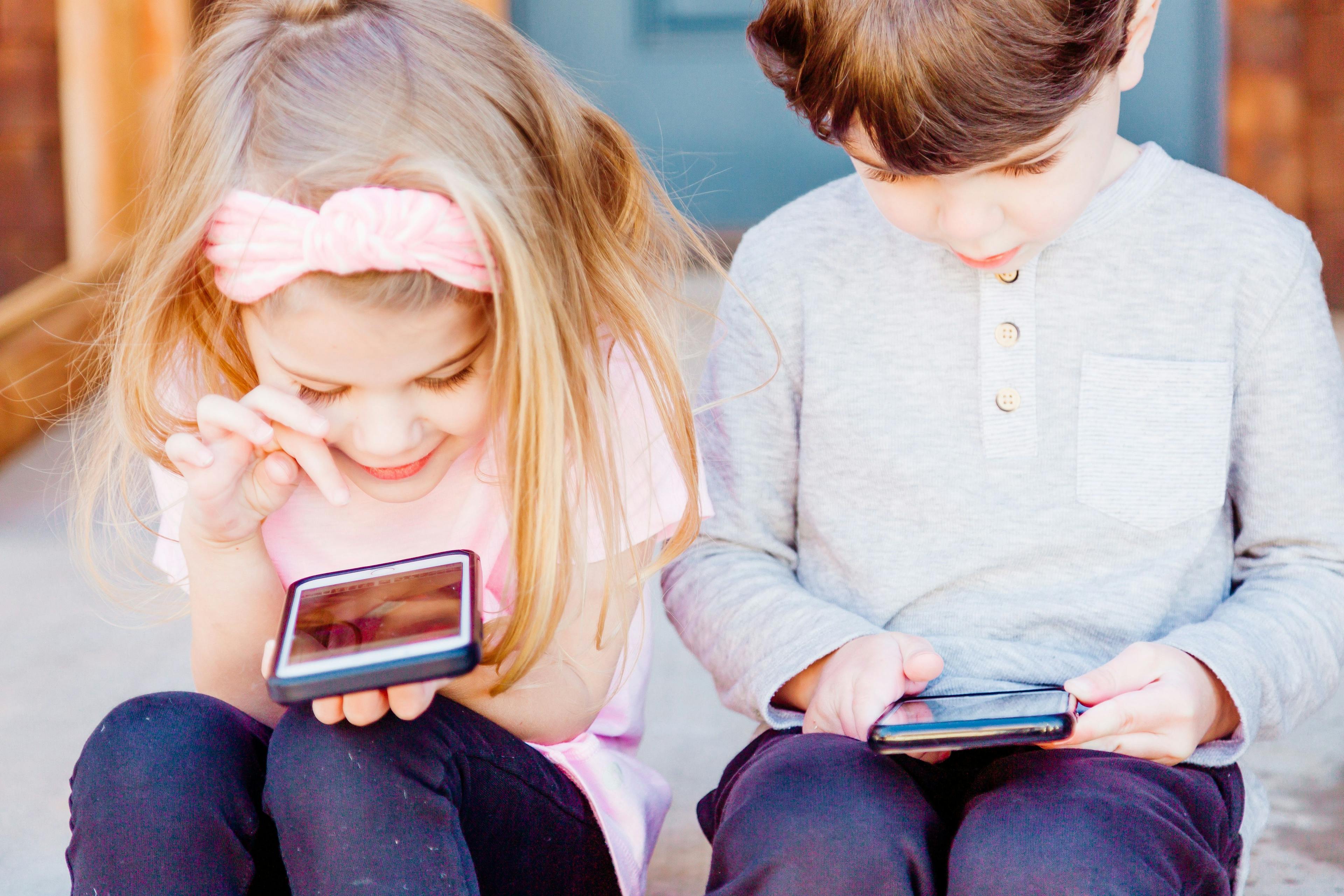 To barn som sitter ved siden av hverandre og driver på hver sin mobiltelefon. De koser seg.